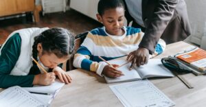Primary school educator with teacher background check helping student with worksheet at desk