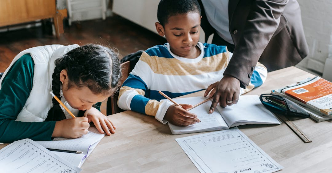 Primary school educator with teacher background check helping student with worksheet at desk